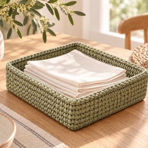 Pode incluir: Uma cesta quadrada de crochê na cor verde sálvia com uma trama de fio espessa e texturizada. Dentro da cesta, há uma pilha de guardanapos de tecido branco dobrados. A cesta possui um design estruturado e baixo com lados verticais.