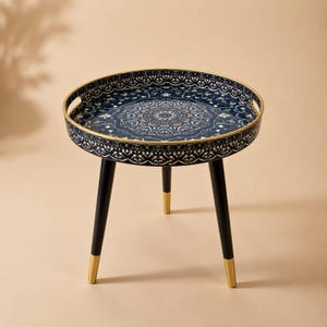 May include: A round side table featuring a dark blue tray top with a white and gold mandala pattern and a gold rim. The table stands on three black legs that have gold-colored tips at the bottom, providing a stable base for the circular surface.