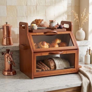 May include: A two-tier wooden bread box with clear glass front panels and handles. The top shelf holds a loaf of bread on a wooden board, while the bottom shelf contains sliced bread. A copper coffee maker and a white vase are nearby.