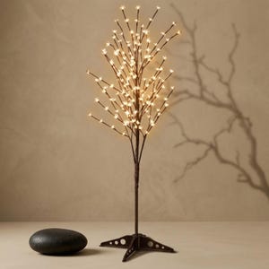 May include: A decorative LED tree with thin, dark brown branches tipped with small, glowing warm white flower-shaped lights. The tree stands on a dark brown four-legged metal base and is accompanied by a smooth, dark gray oval stone.