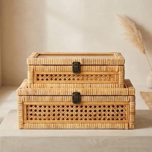 May include: Two rectangular storage boxes made of light brown woven rattan and cane webbing. Each box features a hinged lid with a clear top panel, a dark metal latch closure, and a decorative open-weave cane pattern on the front side.