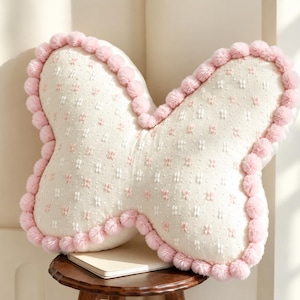 May include: A butterfly-shaped throw pillow in cream fabric with a textured pattern of small pink and white stitched dots. The edges of the pillow are trimmed with a continuous border of soft, round, light pink pom-poms.