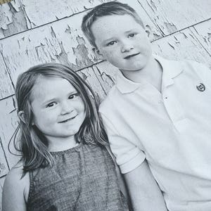 May include: A black and white photograph of a young girl and boy standing in front of a weathered wooden wall. The girl is wearing a sleeveless denim dress and the boy is wearing a white collared shirt with a logo on the pocket.