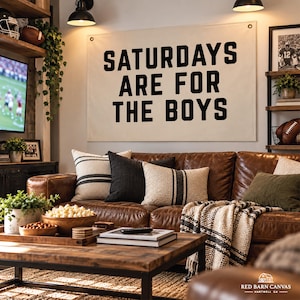 Puede incluir: Un gran tapiz rectangular de lona color hueso con el texto negro SATURDAYS ARE FOR THE BOYS en una fuente sans-serif negrita. El cartel está montado con ojales metálicos en las esquinas superiores sobre una pared.