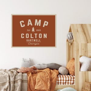 May include: A rust-colored wall banner with the word "CAMP" in white block letters, along with "EST 2023" and "COLTON HARTWELL Georgia". The banner is hanging above a bed with orange bedding and decorative pillows. A wooden house-shaped bed frame is on the right.