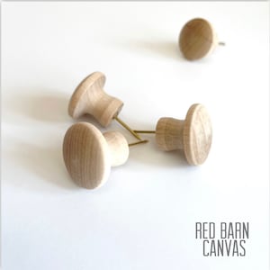 May include: Four wooden knobs with a mushroom shape and a small metal nail protruding from the top. The knobs are light brown in color and are arranged on a white surface.
