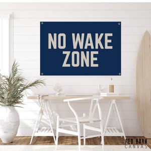 May include: A navy blue banner with the words "NO WAKE ZONE" in large, white block letters. The banner is hanging on a white wall above a desk with a white chair and a surfboard. The banner has grommets in each corner.