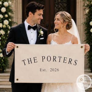Puede incluir: Un estandarte rectangular de lona color hueso con el texto impreso en negro THE PORTERS y Est. 2026. El estandarte incluye cuatro ojales metálicos de color latón, uno en cada esquina, y es sostenido horizontalmente por dos personas con ropa de boda.