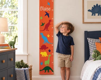 May include: An orange canvas growth chart hanging on a wall, featuring the name Blaise, colorful dinosaur illustrations, and measurement markings in inches. The vertical banner is suspended from a wooden rod with a rope for wall mounting.