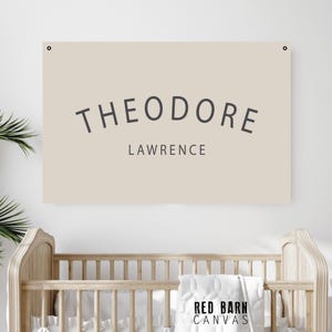 May include: A beige canvas sign with the name "THEODORE" arched above "LAWRENCE" in dark gray letters. The sign is mounted on a white wall above a wooden crib. The Red Barn Canvas logo is visible on a blanket.