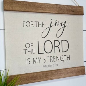 May include: A beige canvas wall hanging with a wooden frame. The canvas features black text that reads "For the joy of the Lord is my strength, Nehemiah 8:10".