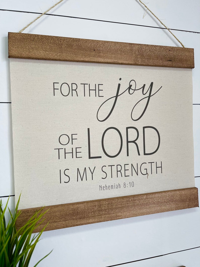 May include: A beige canvas wall hanging with a wooden frame. The canvas features black text that reads "For the joy of the Lord is my strength, Nehemiah 8:10".