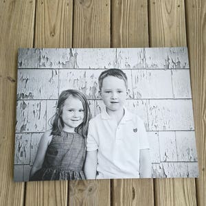 May include: Black and white photograph of two children standing in front of a weathered white wall. The girl is wearing a sleeveless dress and the boy is wearing a white polo shirt.