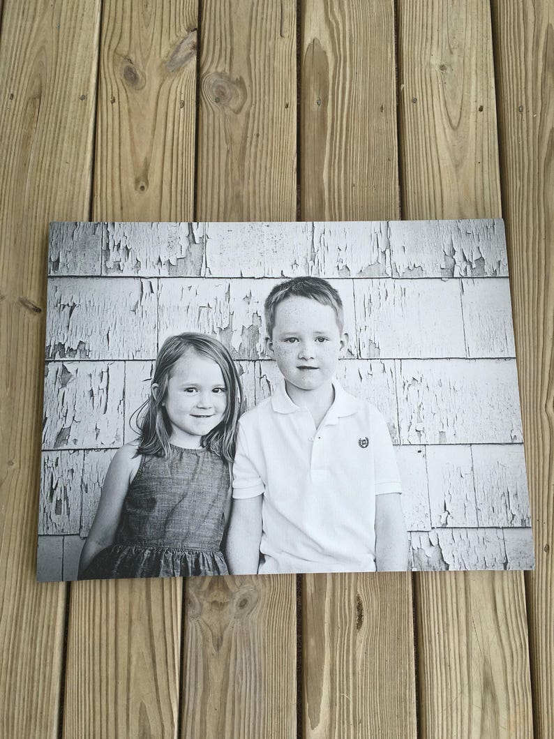 May include: Black and white photograph of two children standing in front of a weathered white wall. The girl is wearing a sleeveless dress and the boy is wearing a white polo shirt.