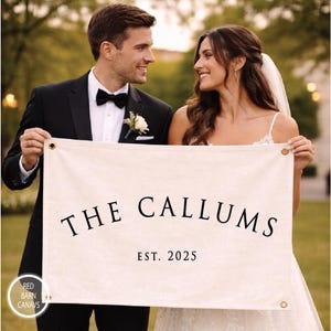 May include: A couple holds a cream-colored banner with the text "THE CALLUMS EST. 2025" in black. The banner has gold grommets. The man wears a black tuxedo and bow tie, and the woman wears a white wedding dress.