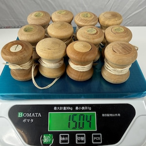 May include: A collection of twelve light brown wooden spools, each wrapped with white string and topped with a small circular label, resting on a digital scale that displays a weight of 1504 grams. The scale is white with a green digital screen.
