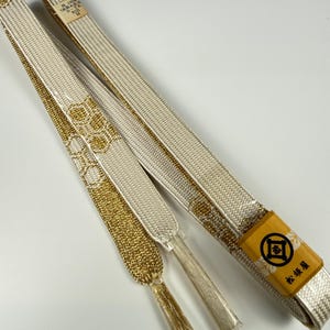 May include: A white and gold obijime cord featuring a woven hexagonal pattern and metallic gold threads. The cord includes a yellow paper tag with a black circular logo and Japanese text, plus a small white label with text including 100%.