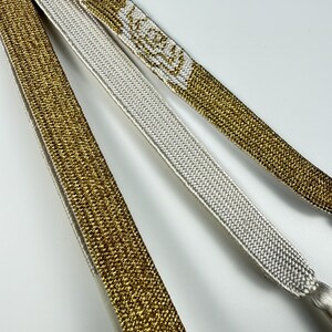 May include: Three flat, woven ribbons arranged in parallel. Two ribbons feature a metallic gold finish with a textured surface, while the central ribbon is a solid, matte white. The gold ribbons display a repeating white floral pattern.