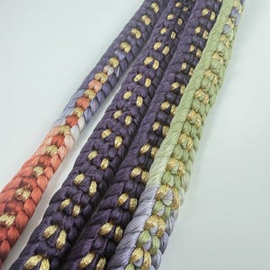 May include: A braided cord featuring a gradient of colors including orange, purple, and light green, interwoven with metallic gold thread. The cord has a thick, textured appearance with a repeating pattern of loops and metallic accents throughout.
