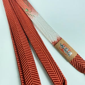 May include: A long, braided cord featuring a deep rust-orange color with a prominent herringbone pattern. One section transitions into a white segment with subtle green accents. A small, rectangular tan label with silver Japanese text is attached.