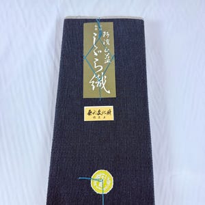 Könnte beinhalten: Ein rechteckiger Stoffballen in Dunkelmarineblau mit einer feinen, vertikal gerippten Textur. Er ist mit einem langen olivgrünen Etikett mit weißem japanischen Text, einem kleineren rechteckigen gelben Etikett und einem runden gelben Aufkleber versehen.