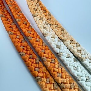 May include: Three braided cords featuring a gradient color transition from deep orange to light peach and white. Each cord is woven with a metallic gold thread interspersed throughout the braided pattern, creating a textured and reflective appearance.