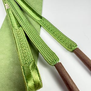 May include: A bright green obijime cord featuring a woven texture with a central decorative pattern and smooth, flat ends. The cord terminates in two brown, cylindrical, felt-like tips. A small white tag with black text is attached to the cord.