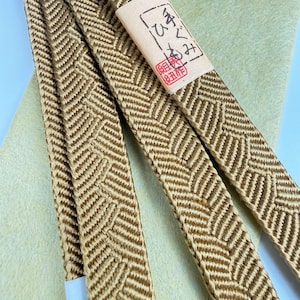 May include: A long, narrow, gold-colored braided cord featuring a repeating chevron pattern. A rectangular paper label is attached to the cord, displaying handwritten Japanese characters and a small red stamp. The cord has a smooth, woven texture.