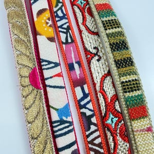 May include: Five long, narrow fabric strips arranged vertically. The first is bright pink with gold embroidery. The second features a white background with blue, yellow, and black floral patterns. The third is orange with a dark blue and white design. The fourth is cream with red and teal embroidery. The fifth is a textured beige with green, red, and black horizontal stripes.