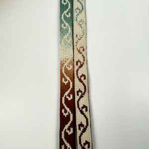 May include: Two long, narrow woven fabric straps featuring a repeating geometric wave pattern. The left strap displays a vertical color gradient transitioning from sage green to rust red. The right strap is cream-colored with a dark red pattern.