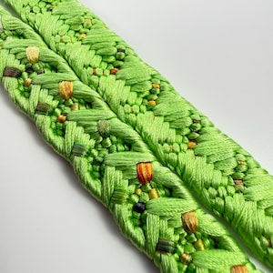 May include: A lime green braided cord featuring a complex woven pattern with integrated accents of yellow, orange, and dark green thread. The textile accessory displays a dense, textured surface created by interlaced fibers in a repeating design.