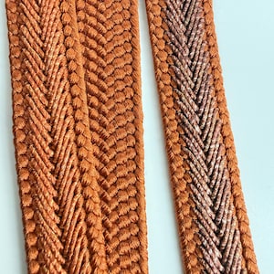 May include: Two strips of burnt orange braided trim featuring a central herringbone pattern. The texture is composed of tightly woven threads with a subtle sheen, creating a raised, dimensional surface that runs vertically along each strip.