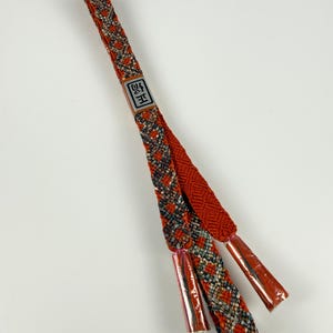 May include: A long, flat, braided cord in vibrant orange, featuring a complex geometric pattern in grey and white. A small rectangular tag with the black Japanese characters 正絹 is attached. The ends are finished with smooth, metallic-toned caps.