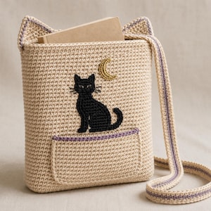 May include: A beige crochet crossbody bag with cat-ear shaped top corners, featuring an embroidered black cat and a gold crescent moon. The bag includes a front pocket with a purple trim, a long matching strap, and a light brown book inside.