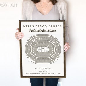 May include: A black and white print of the Wells Fargo Center, home of the Philadelphia Flyers. The print shows a seating chart of the arena with the ice rink in the centre. The text "WELLS FARGO CENTER" and "Philadelphia Flyers" is at the top of the print. The text "CAPACITY: 19,306" and "Est. 1996" is at the bottom of the print. The print is framed in a brown frame.