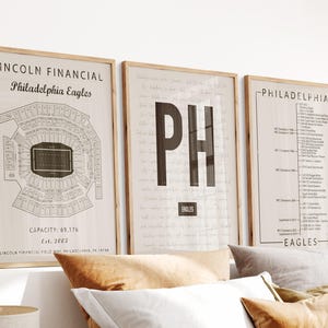 May include: Three framed wall art prints featuring Philadelphia Eagles themes. The left print shows a stadium map, the centre print displays the letters PH, and the right print lists team history and statistics on a light beige background.