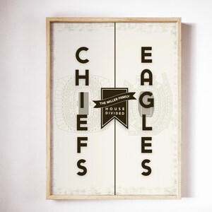 May include: Framed art print featuring the words "CHIEFS" and "EAGLES" in bold black letters. The print includes stadium schematics and a banner that reads "THE MILLER FAMILY HOUSE DIVIDED". The frame is made of light-colored wood.
