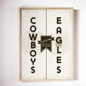 May include: A framed print featuring the words "Cowboys" and "Eagles" in black, bold lettering on a cream backdrop. The design incorporates stadium schematics and a banner stating "The Miller Family House Divided."