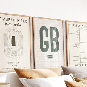 May include: Three framed wall art prints featuring Green Bay Packers themes. The left print shows a Lambeau Field stadium diagram. The centre print displays large green letters GB. The right print features a timeline of Green Bay Packers history.