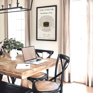 May include: A black framed print of a stadium seating chart with the text "Levi Stadium, San Francisco 49ers, Est. 2014, Capacity: 75,000" hanging on a white wall. A wooden table with a laptop, a mug, and a notebook is in the foreground.