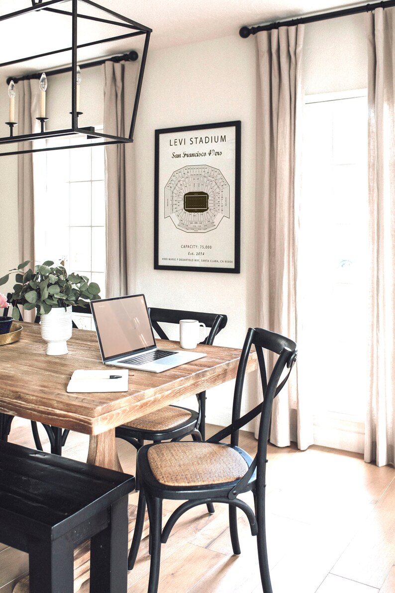 May include: A black framed print of a stadium seating chart with the text "Levi Stadium, San Francisco 49ers, Est. 2014, Capacity: 75,000" hanging on a white wall. A wooden table with a laptop, a mug, and a notebook is in the foreground.