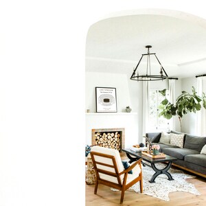 May include: A living room interior with a gray sectional sofa, a white armchair, a coffee table, a fireplace, and a framed print of a stadium seating chart. The room has a white ceiling, white walls, and hardwood floors.
