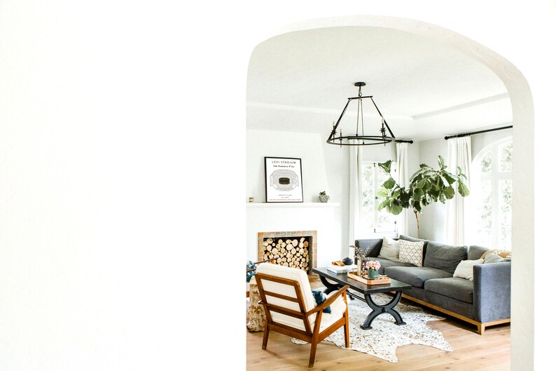 May include: A living room interior with a gray sectional sofa, a white armchair, a coffee table, a fireplace, and a framed print of a stadium seating chart. The room has a white ceiling, white walls, and hardwood floors.