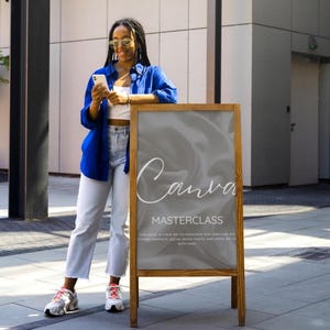 May include: A wooden A-frame sidewalk sign featuring a grey satin-textured background with the white text "Canva MASTERCLASS" and smaller descriptive text below. The sign is displayed outdoors on a paved surface near a modern building exterior.