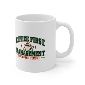 Puede incluir: Una taza de cerámica blanca con un diseño gráfico que presenta el texto COFFEE FIRST MANAGEMENT DECISIONS SECOND en letras de color verde oscuro y rojo, junto a una ilustración de una taza de café humeante y pequeñas estrellas rojas.