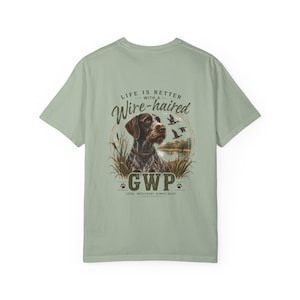 May include: A sage green t-shirt featuring a graphic on the back of a German Wirehaired Pointer dog in tall grass near a lake. The text reads: LIFE IS BETTER WITH A Wire-haired GWP LOYAL, INTELLIGENT, ALWAYS READY, with silhouettes of flying ducks.