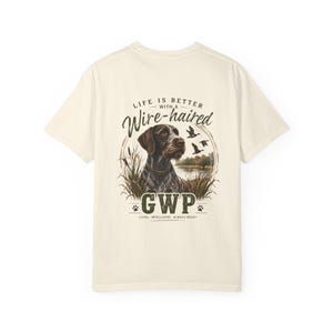 May include: A cream-coloured t-shirt featuring a graphic on the back of a German Wirehaired Pointer dog in tall grass near a lake with flying ducks. The text reads: LIFE IS BETTER WITH A Wire-haired GWP LOYAL. INTELLIGENT. ALWAYS READY.