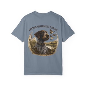 Peut inclure: Un t-shirt bleu ardoise avec un motif au dos représentant un chien braque allemand à poil dur portant un collier, entouré de hautes herbes et de canards en vol, avec le texte GERMAN WIREHAIRED POINTER arqué au-dessus de l'illustration.