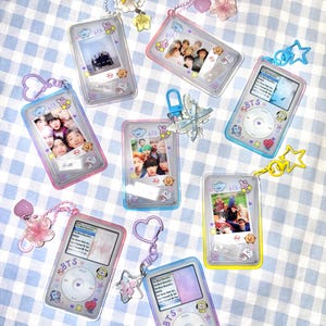 May include: A collection of seven rectangular acrylic keychains designed to resemble portable music players and photo frames. Each features colorful borders, small cartoon character stickers, and attached metal clasps with charms like stars, hearts, and flowers.