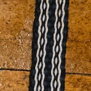 May include: A long, narrow woven band featuring a repeating geometric pattern in dark navy blue and white threads. The textile strip is constructed with a series of vertical, interlocking diamond shapes and terminates at both ends in loose, unknotted fringe.